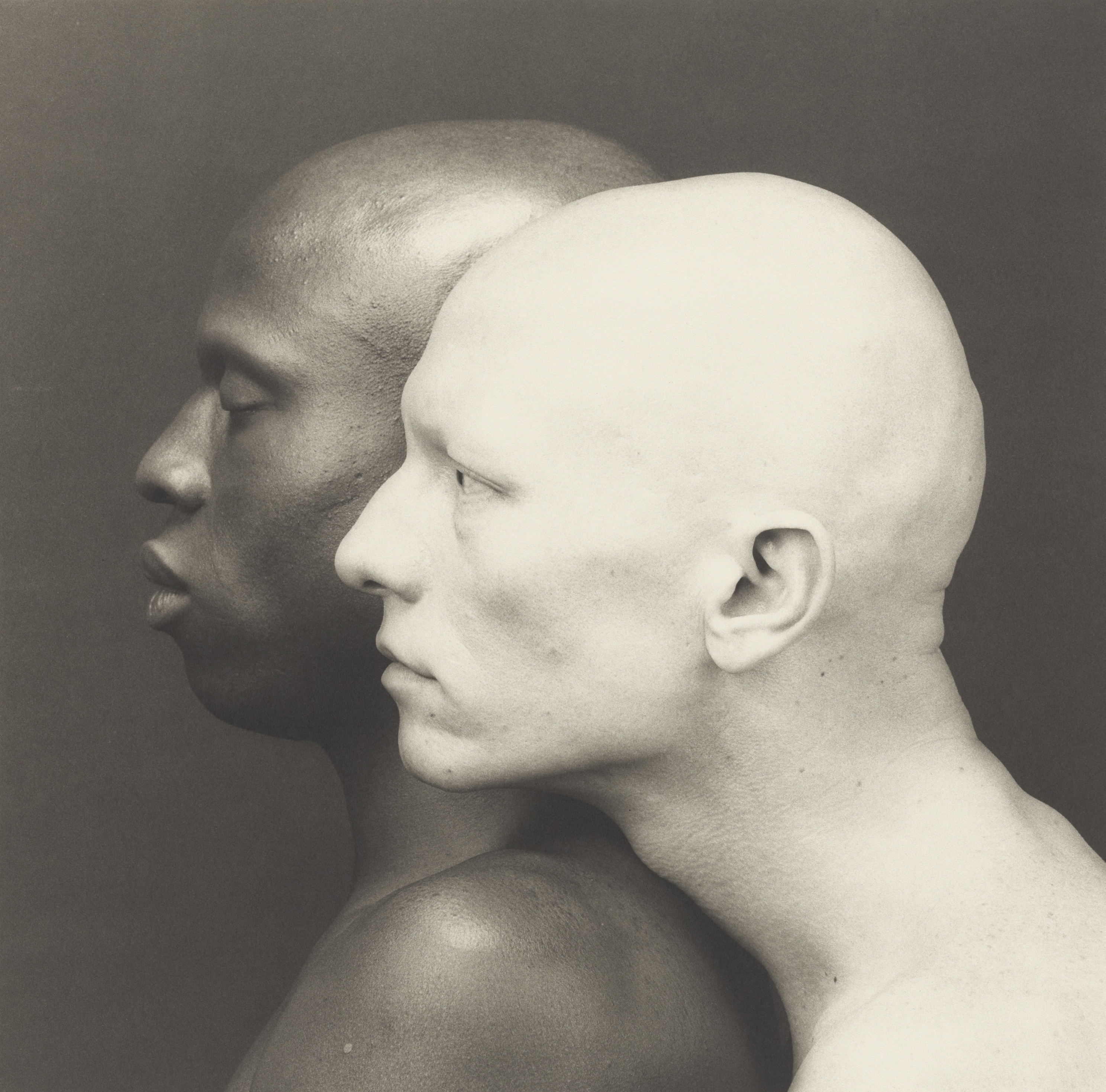 Black and white portrait of two bald men standing profile against a studio backdrop.