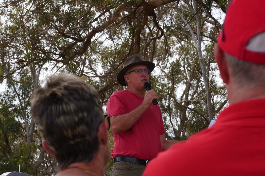 A man in a red shirt and akubra hat speaks at a microphone in front of a big tree.