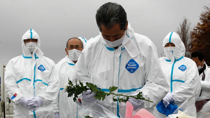 Evacuees dressed in protective suits offer flowers and prayers for victims of Japan's 2011 disaster.