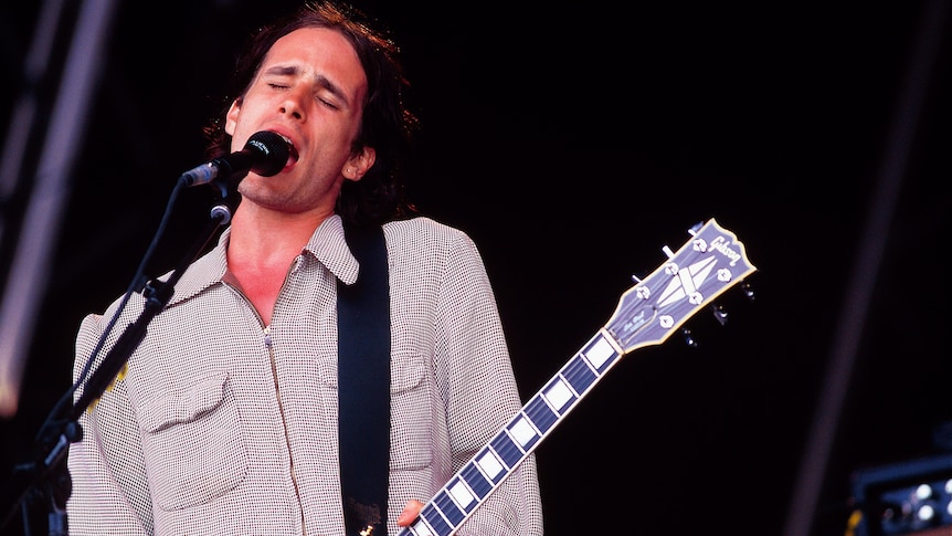Jeff Buckley sings and plays guitar onstage. He wears a grey button-up shirt.