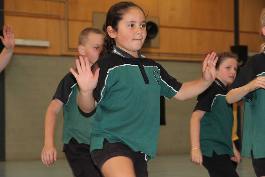A young girl in a primary school uniform dances with her hands up in the air