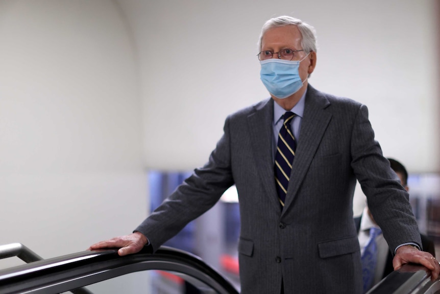 A man with white hair wearing a mask holds his hands out on either side of an escalator.