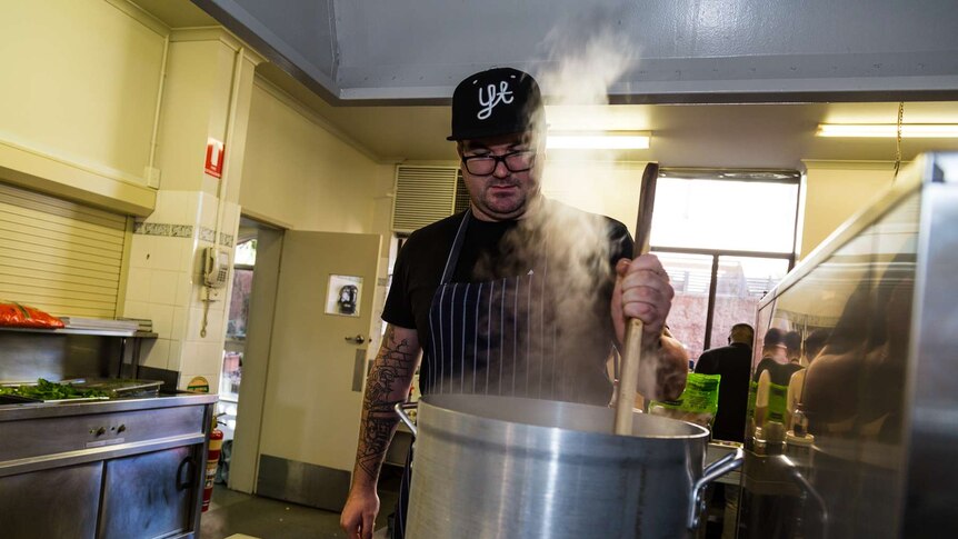 A man in a hat stirs a pot of food in an industrial kitchen.