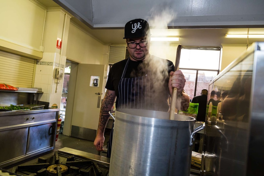 A man in a hat stirs a pot of food in an industrial kitchen.