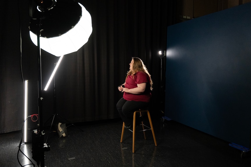 A young woman sits on a stool against a black background with a large studio light shining on her