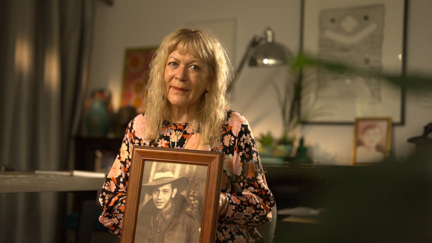 a blonde woman in a floral dress holding a photo of her father