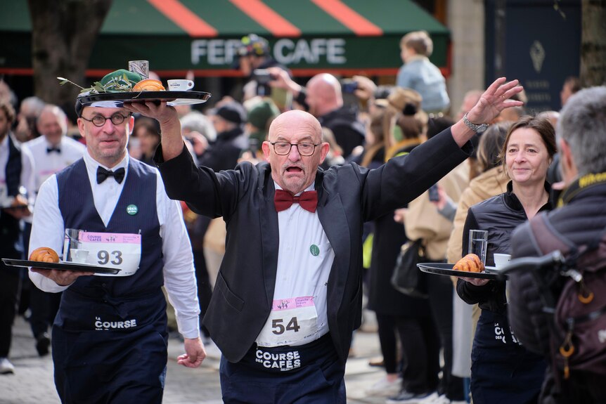 A bald man with glasses holds his arms up as he races other waiters.