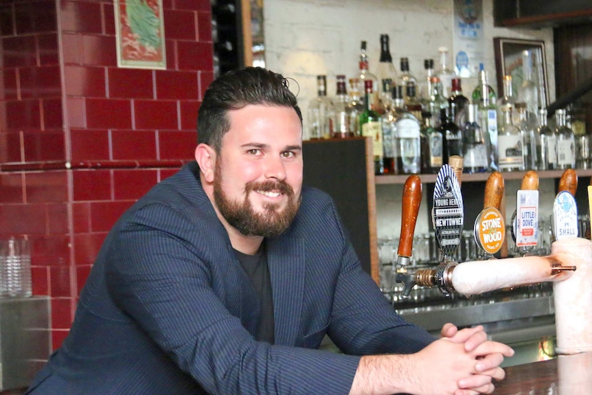 Man in blue jacket sitting at the bar in a pub with bottles behind him