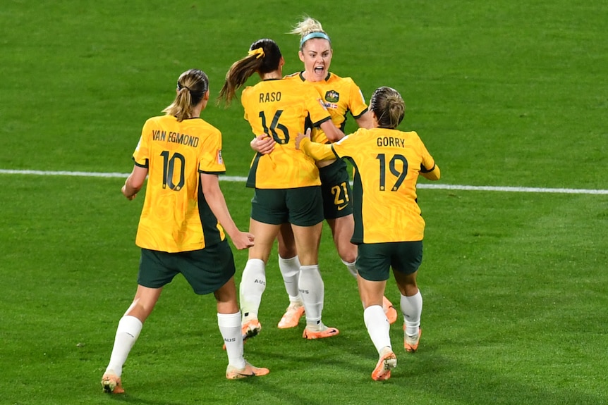 A gorup of four female soccer players in green and gold jerseys hug each other in celebration.