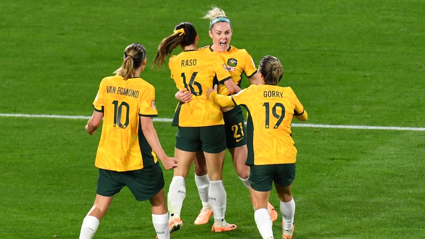A gorup of four female soccer players in green and gold jerseys hug each other in celebration.