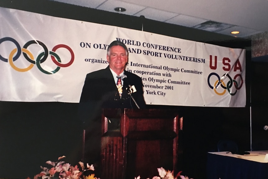 Laurie Smith standing at a podium in a black suit. A banner for the conference is behind him.