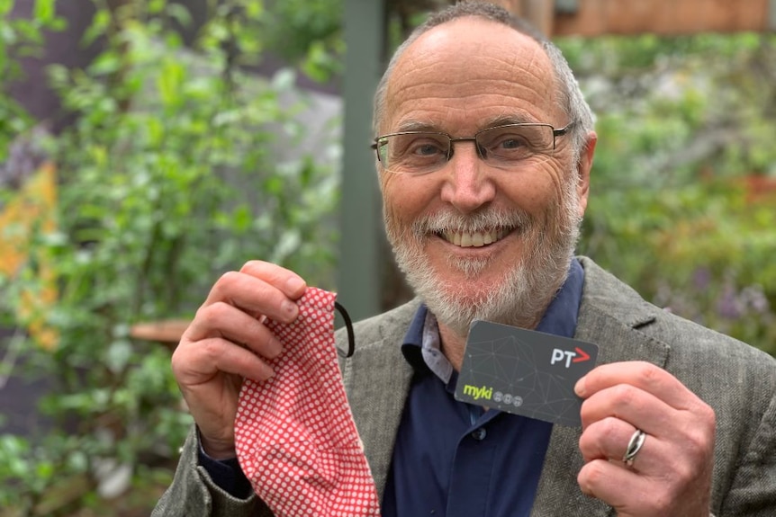 A man with a short grey beard stands in a garden holding a face mask and a Myki public transport card.