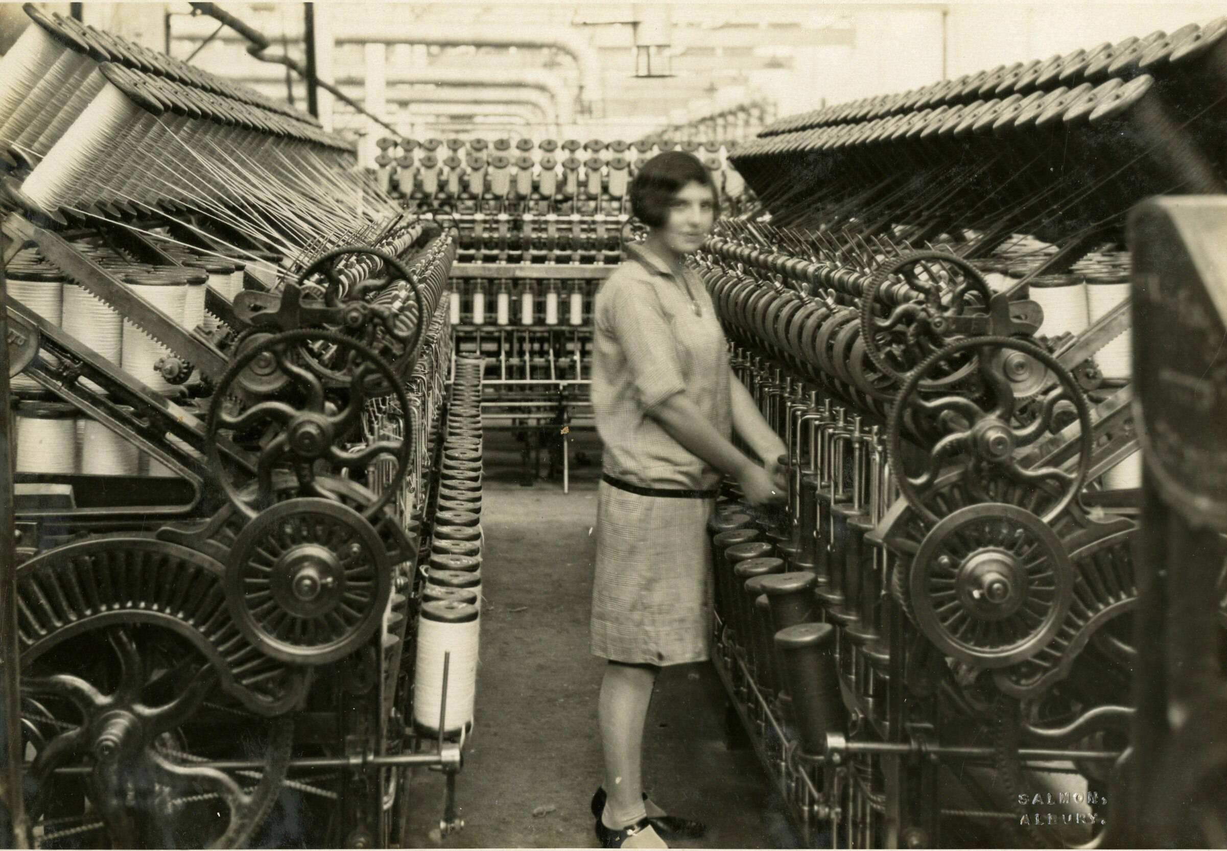 Old sepia image of woman standing in front of a row of wool spinning machines.