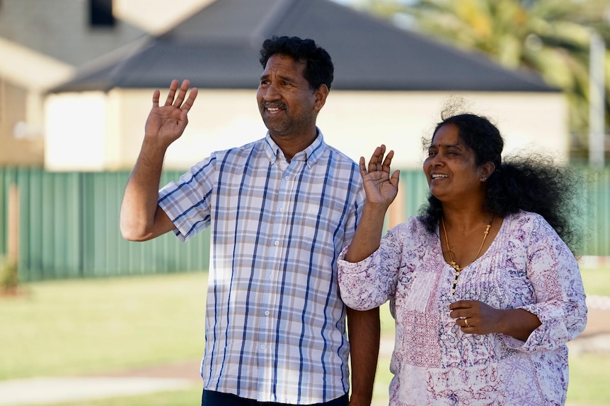 A man and woman wave while standing in a park