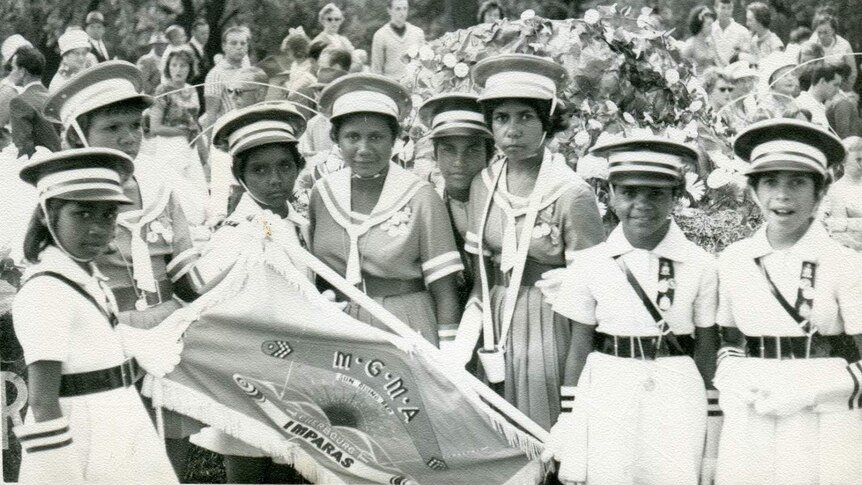 A black and white photo of Indigenous girls holding a flag as part of the Cherbourg marching girls in Melbourne 1962.