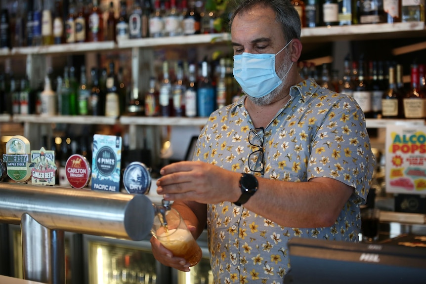 A man with a greying beard and blue collared shirt pours a beer behind a bar
