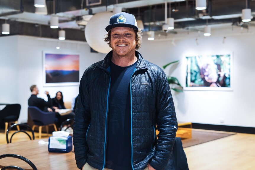 A man wearing a hat and puffer jacket smiles inside an art gallery
