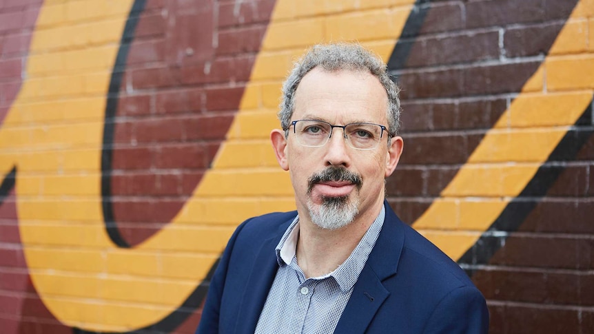 Portrait of Nick Gadd, pictured alongside a brick wall with yellow letters