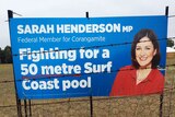 A blue election sign with a woman's head on it. The sign promises a pool and stands in a field with barbed wire in front.