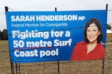 A blue election sign with a woman's head on it. The sign promises a pool and stands in a field with barbed wire in front.