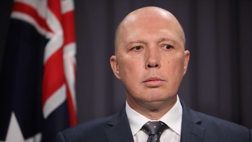 Tight shot of Dutton standing in front of an Australian flag. He is looking down towards a journalist (not in frame).