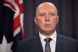 Tight shot of Dutton standing in front of an Australian flag. He is looking down towards a journalist (not in frame).
