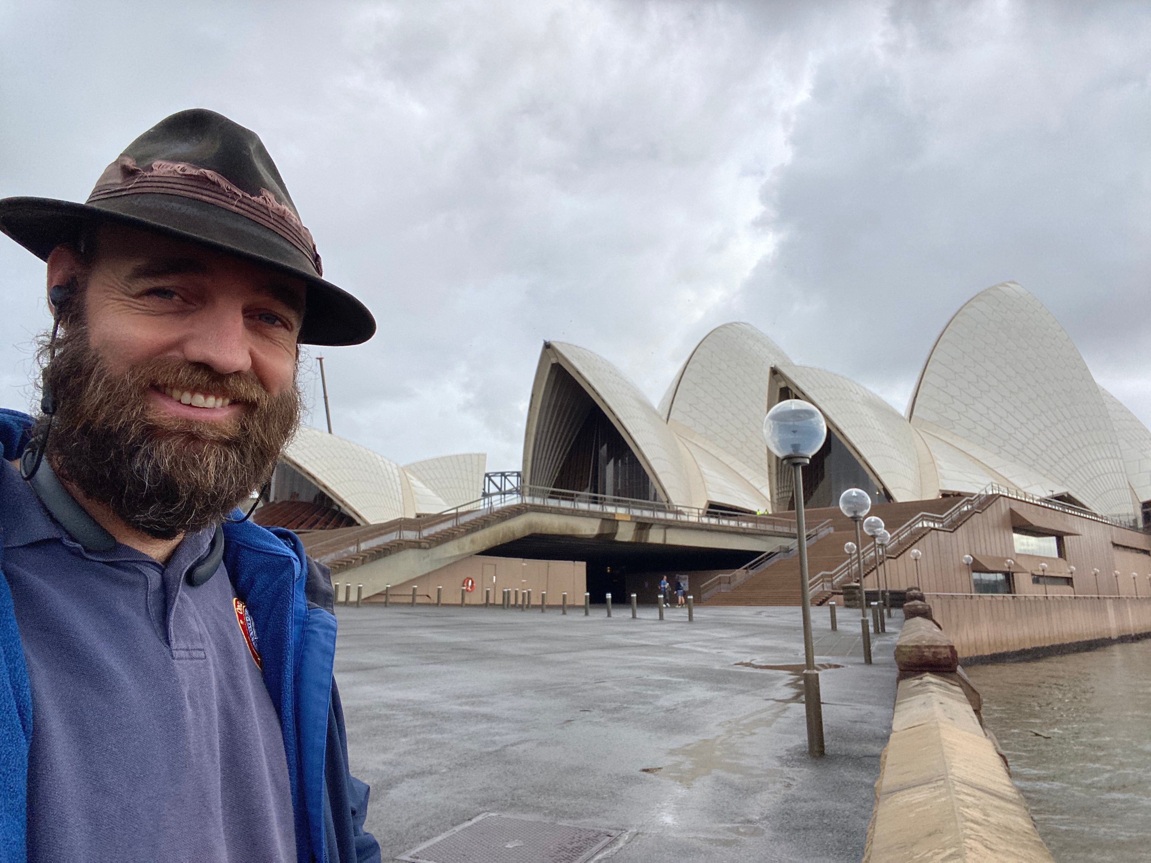A man in hat smiles in front of the Sydney Opera House on a cloudy day 