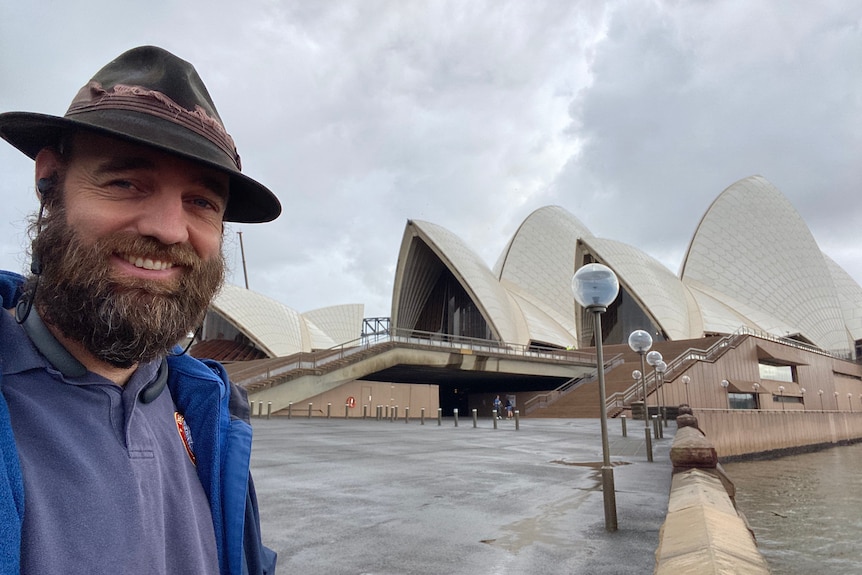 A man in hat smiles in front of the Sydney Opera House on a cloudy day 