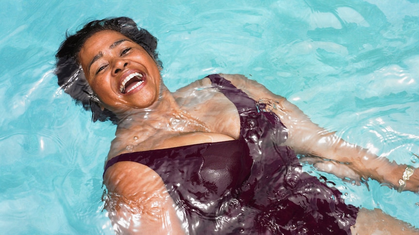 Older woman in bathing suit smiling widely as she floats at the surface of a swimming pool's water.