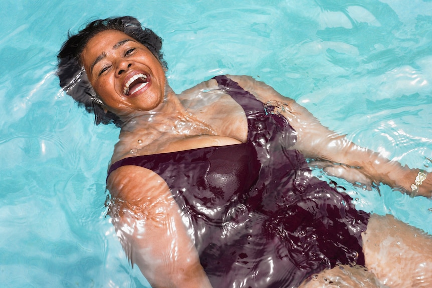 Older woman in bathing suit smiling widely as she floats at the surface of a swimming pool's water.