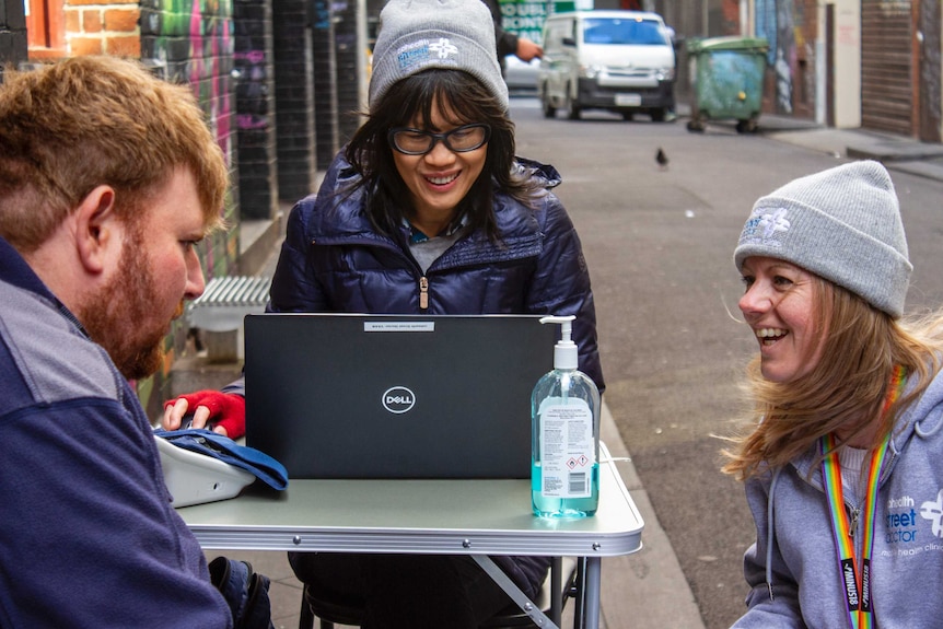 Two women and a man sit around a fold-up table in an alleyway.