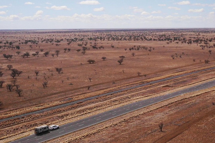 An aerial view of an outback road with red earth surrounding and blue sky with a single vehicle on the road