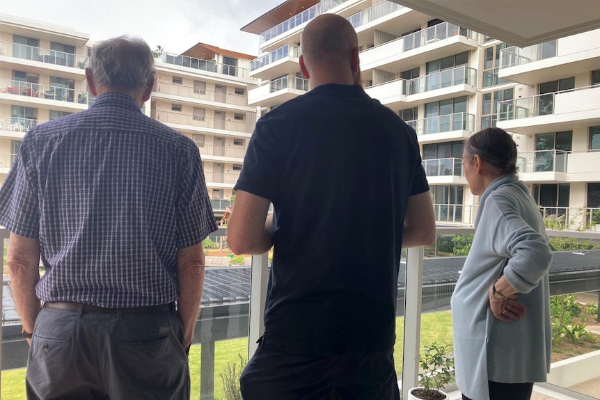 Two men and a woman stand on an apartment building balcony, their backs to the camera