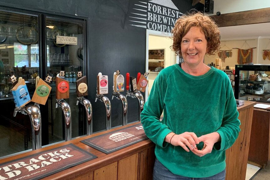 A woman with red curly hair and a green jumper stands at a bar beside eight beer taps.