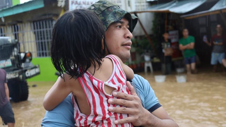 A rescuer in a hat carries a child to safer grounds as floodwaters rise. 