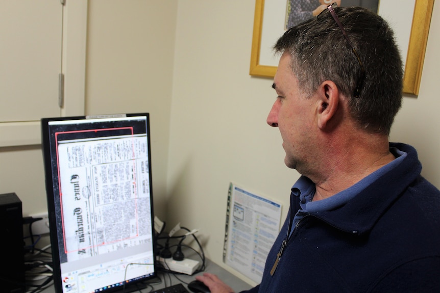 A man looks at an old newspaper on a computer monitor. 