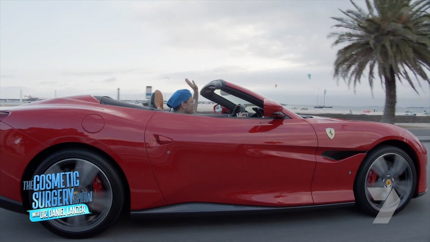 Dr Lanzer waves from a Ferrari sports car as he drives it past a palm tree.