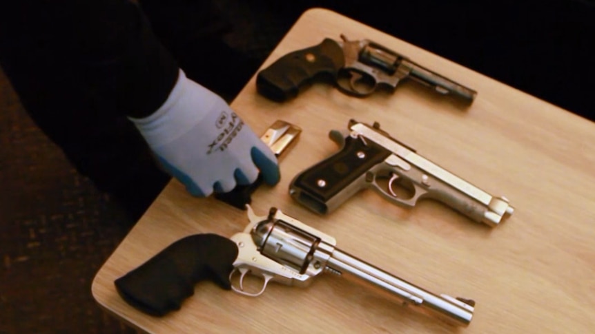 A small table with three guns on it, two revolvers and a handgun. A gloved hand is placing a clip of ammunition next to them.