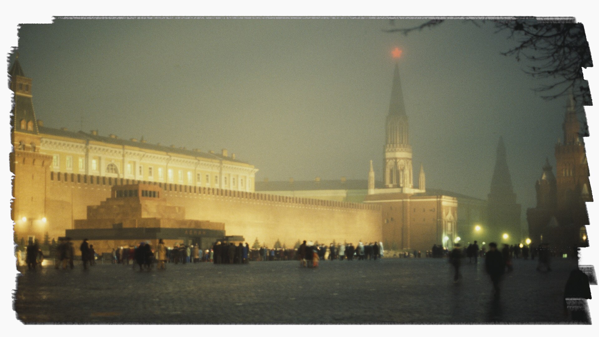 People walk past a large illuminated walled building on a public square. It is night and there appears to be fog.