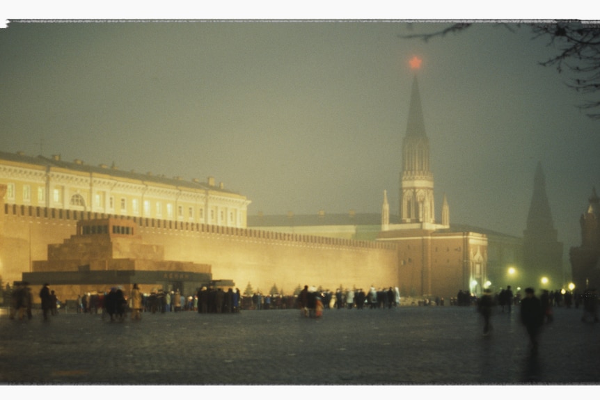 People walk past a large illuminated walled building on a public square. It is night and there appears to be fog.