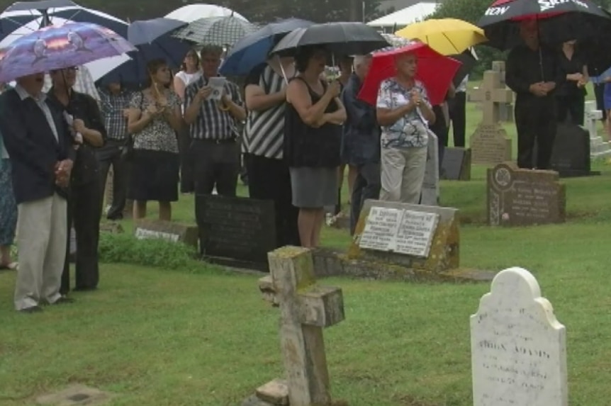 Mourners stand in the rain under umbrellas at Colleen McCullough's funeral.