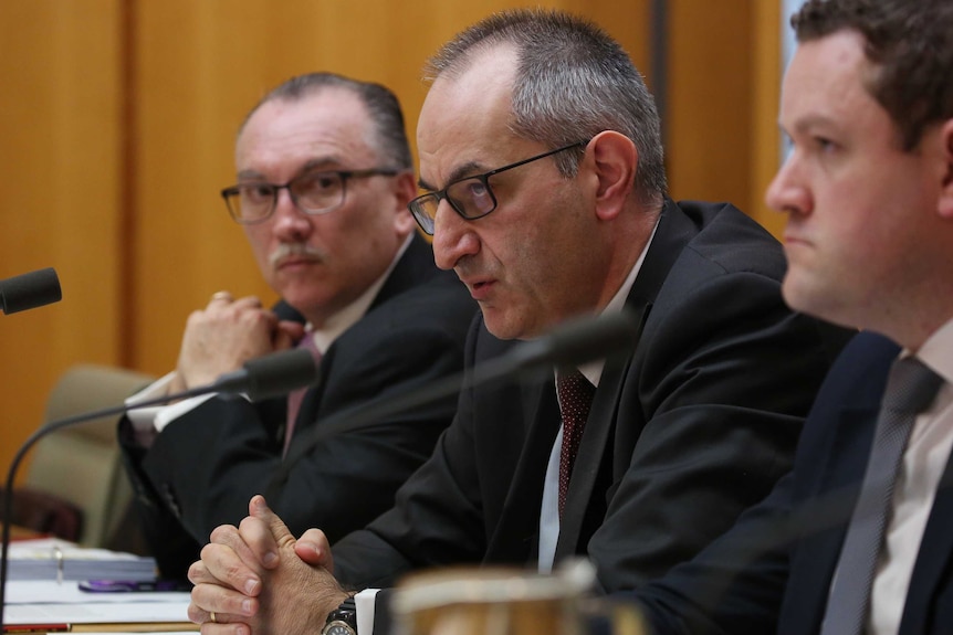 Mr Pezzullo is flanked by Home Affairs officials, sitting behind a microphone. He's wearing black rimmed glasses.