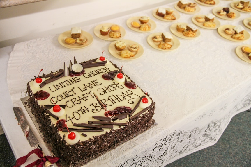 A massive birthday cake sits on a table beside plates of morning tea. 