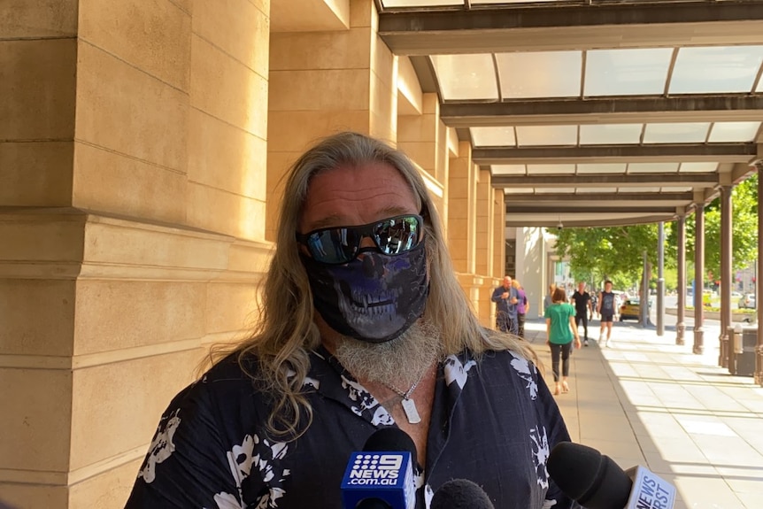 A man with long grey hair in a Hawaiian shirt standing outside court buildings