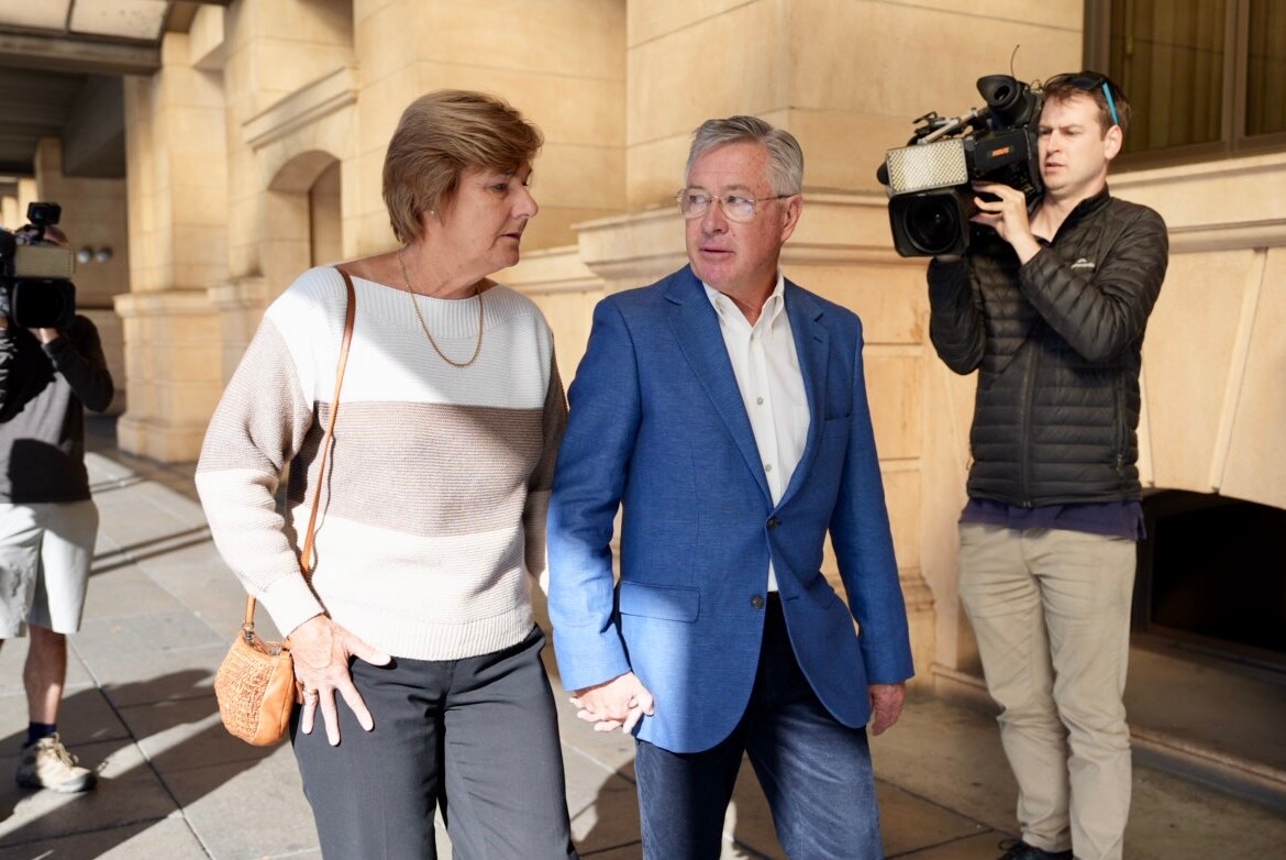 A man and a woman hold hands as a cameraman films them walking outside court