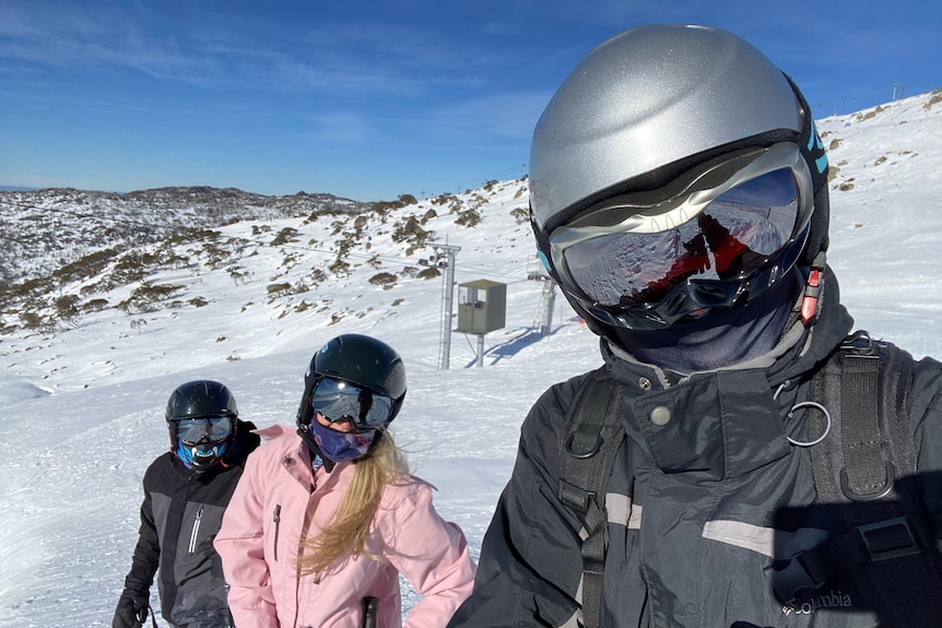 A man and two children wearing ski masks, goggles and helmets face the camera. Snow fields are visible in the background.