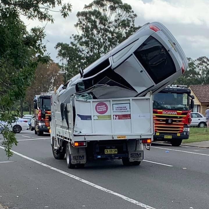 A white car sits upside down in a truck trailer in the middle of a road.