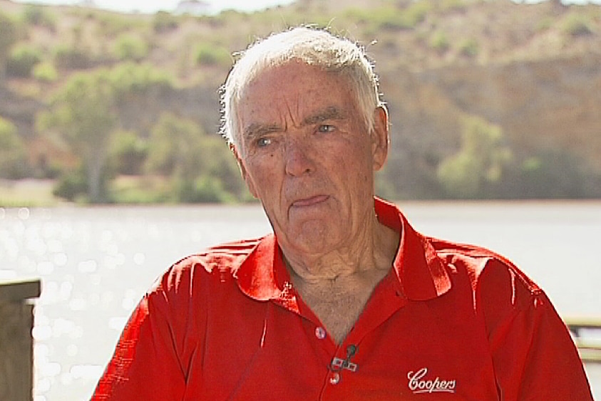 An elderly man wearing a red polo shirt in front of a river with cliffs