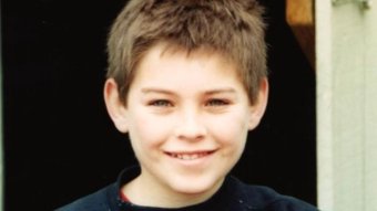 A close up photo of a young boy, smiling, with short cropped brown hair.
