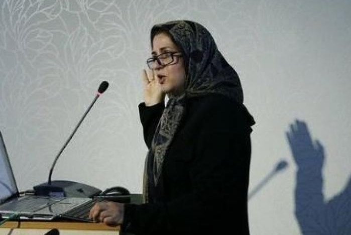 Meimanat Hosseini Chavoshi speaks at a lectern, with her shadow visible on the wall behind.